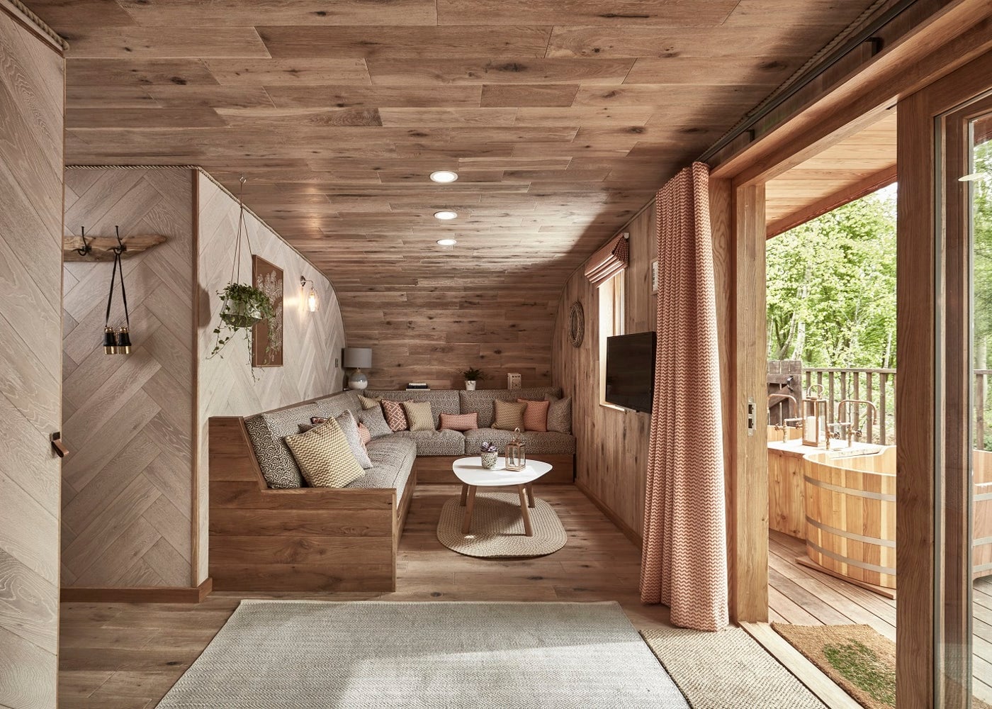 Cozy wood-paneled living room with an L-shaped sofa, round coffee table, and a TV, opening to a wooden deck with a hot tub outside.