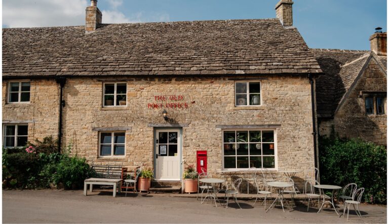 Stone cottage with 'The Old Post Office' sign, a red postbox outside, and white metal café tables and chairs on a sunny day.
