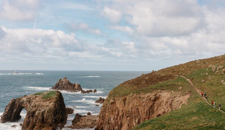 Hikers walk a narrow grassy cliff path along the coast, with the sea to the left and rugged rocks below; a lighthouse is visible offshore in the distance.