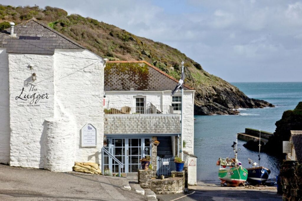 Whitewashed hotel-restaurant The Lugger beside a small harbor, with rocky hillside and boats moored in calm water nearby.