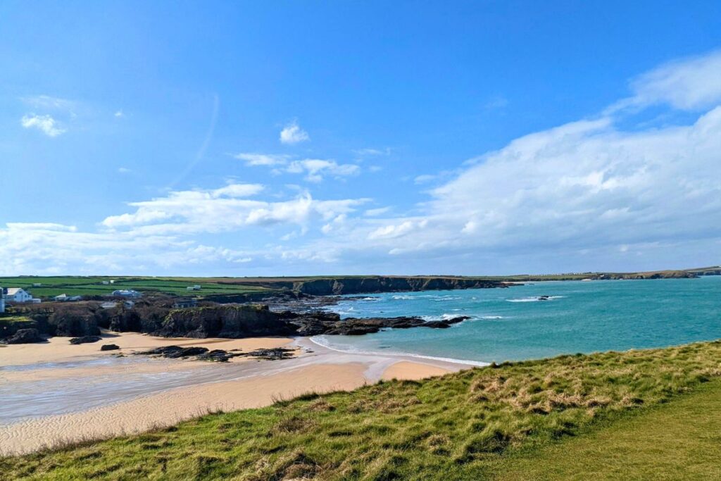 Sunny coastal scene with sandy beach, rocky shore, turquoise sea, and a grassy foreground hill.