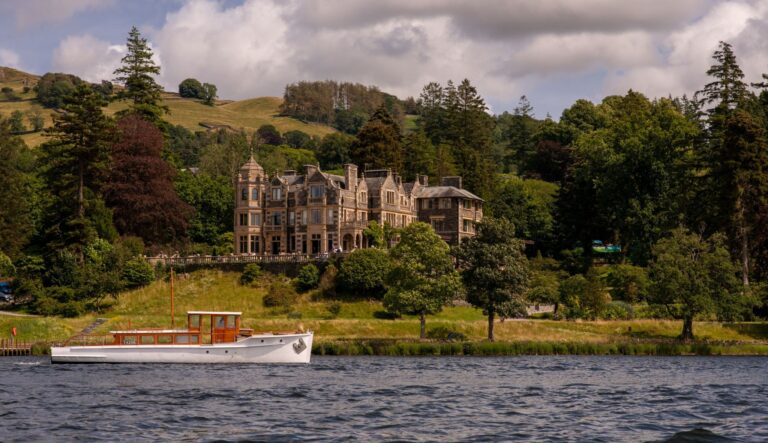 Stone mansion with ornate towers along a riverbank, surrounded by trees and hills; a white boat rests at the water's edge in the foreground.