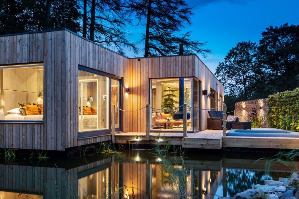 Modern wooden villa on a pond at dusk, with large glass walls and a lit, inviting interior. A wooden deck extends over the water.