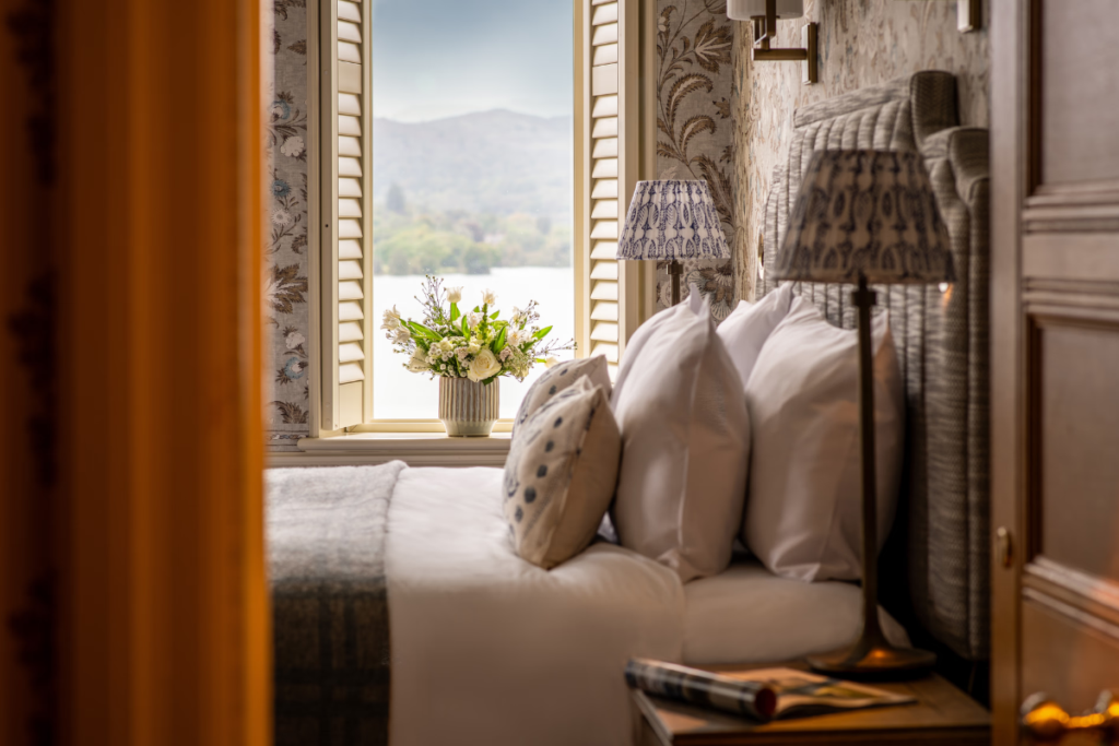 Cozy hotel bedroom nook with a window seat, white bedding, patterned pillows, and two lamps; a vase of white flowers sits on the windowsill with a mountain view outside.