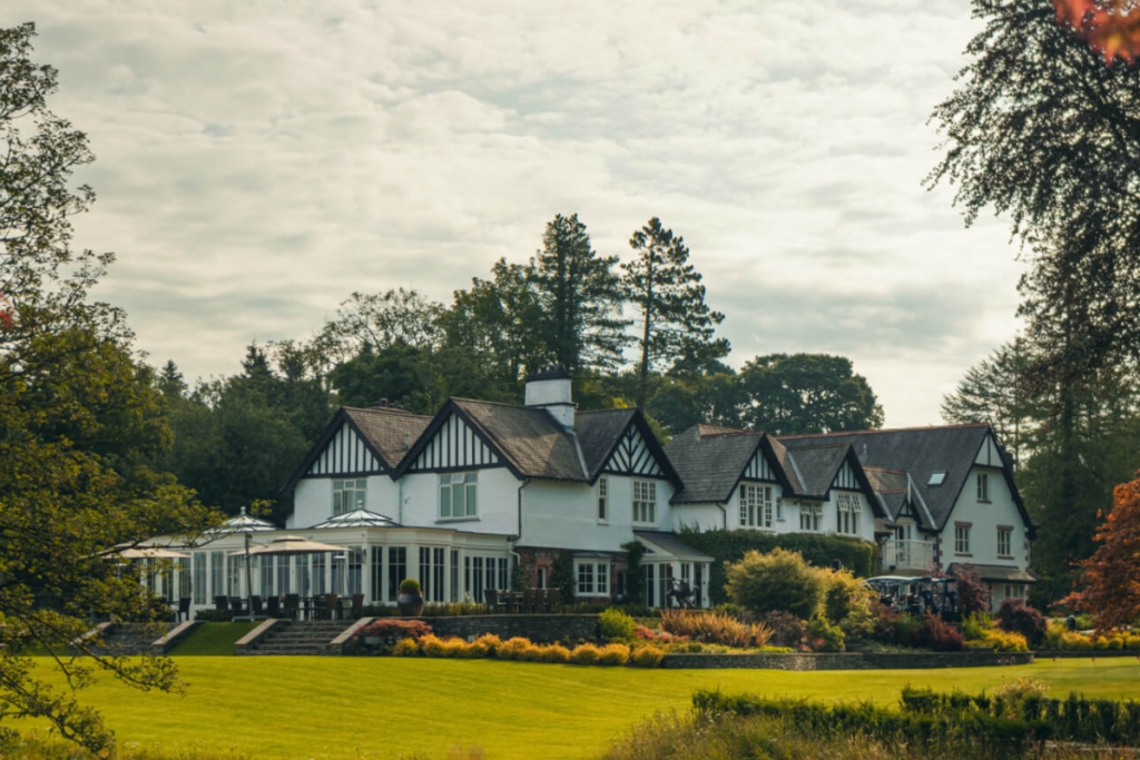 Large white Tudor-style country house with dark timber framing, surrounded by manicured gardens and a sunny lawn.
