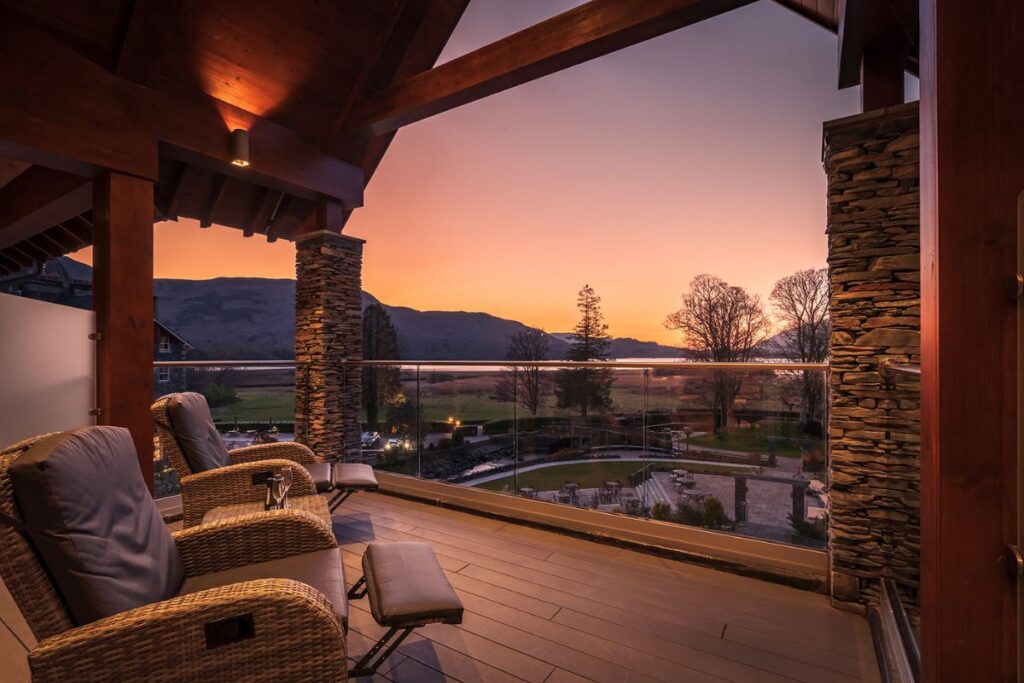 Cozy covered balcony with wicker lounge chairs and stone columns, overlooking a sunset-lit valley and rolling hills.