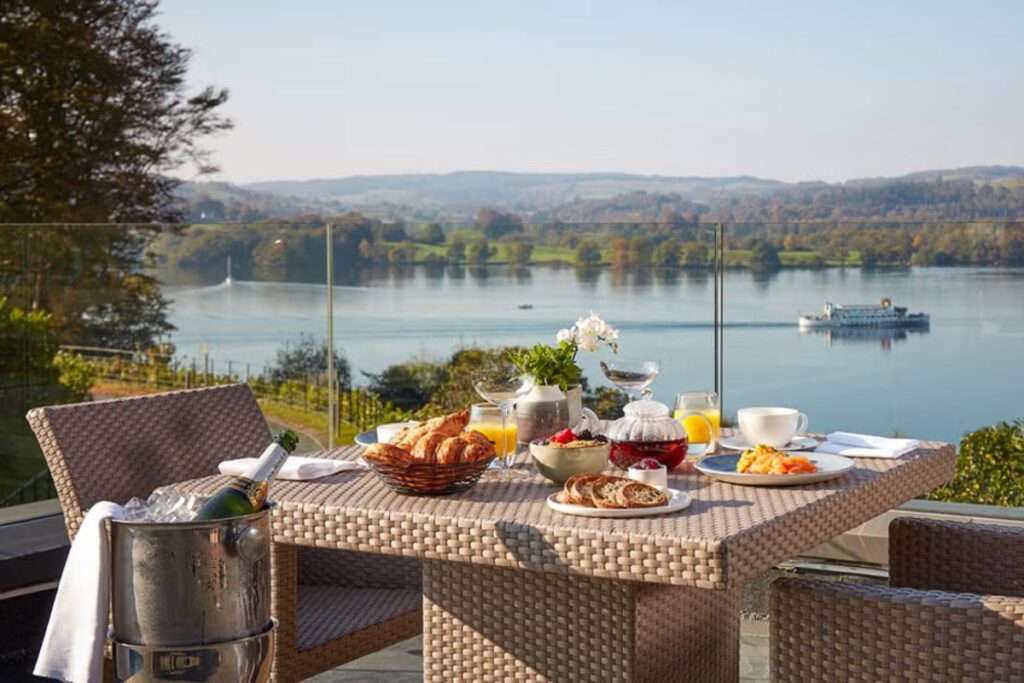 Outdoor wicker dining table set for breakfast on a terrace, with croissants, juice, fruit, and a bottle chilling in a cooler, overlooking a calm lake and distant hills.